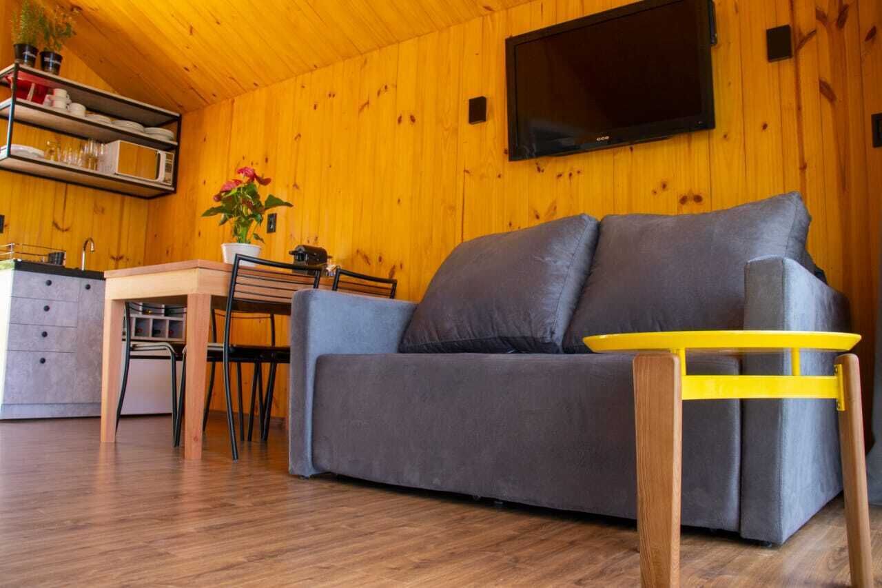 Interior acolhedor de uma cabana com um sofá cinzento, mesa de jantar de madeira e uma TV fixada na parede.