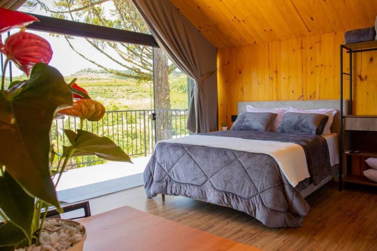 A bedroom with a bed, wooden walls, a balcony with plants, and a view of the outdoors.