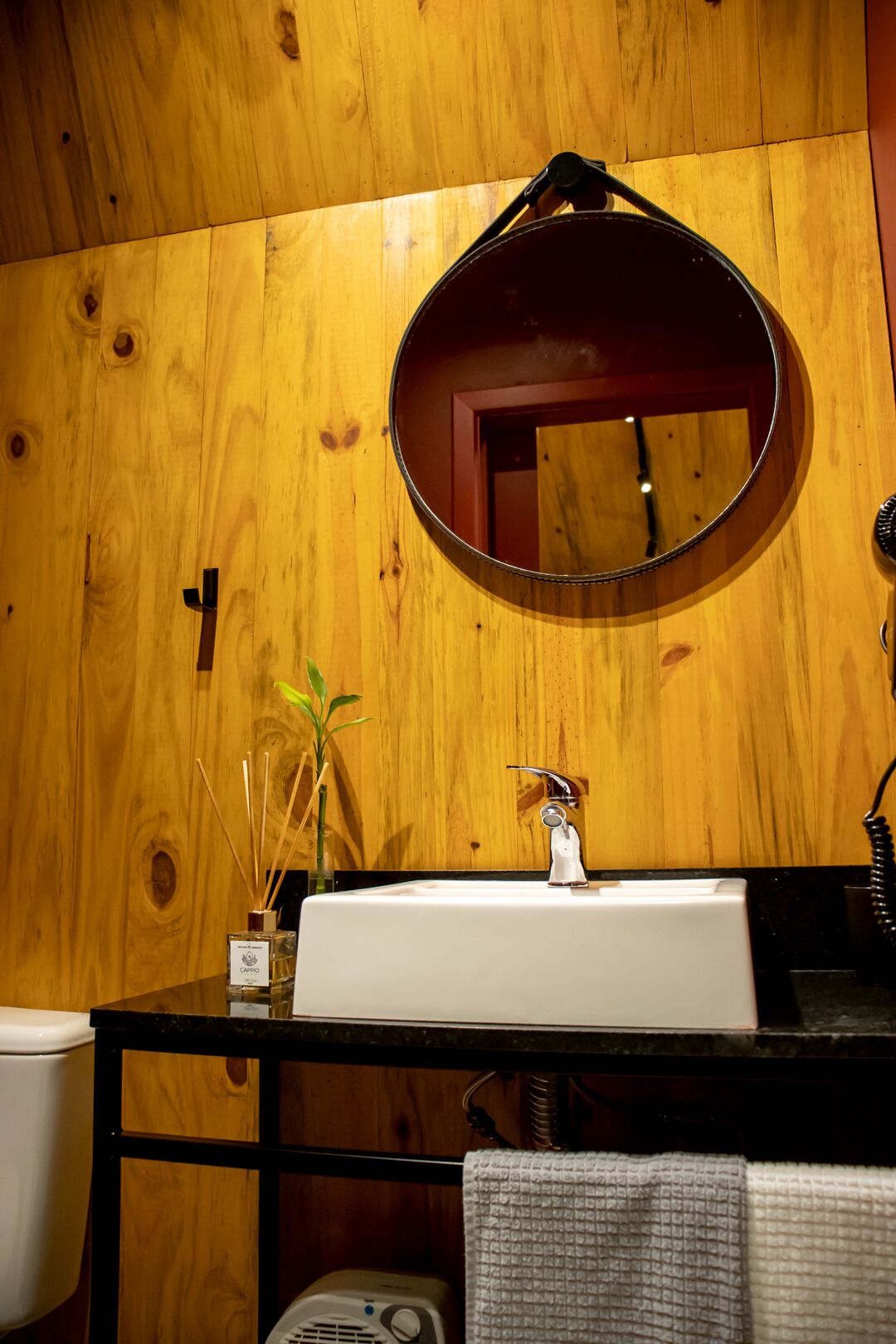 Een badkamer met een houten wand, een ronde spiegel, een wit wastafelmeubel en een handdouche, inclusief een handdoekhouder.