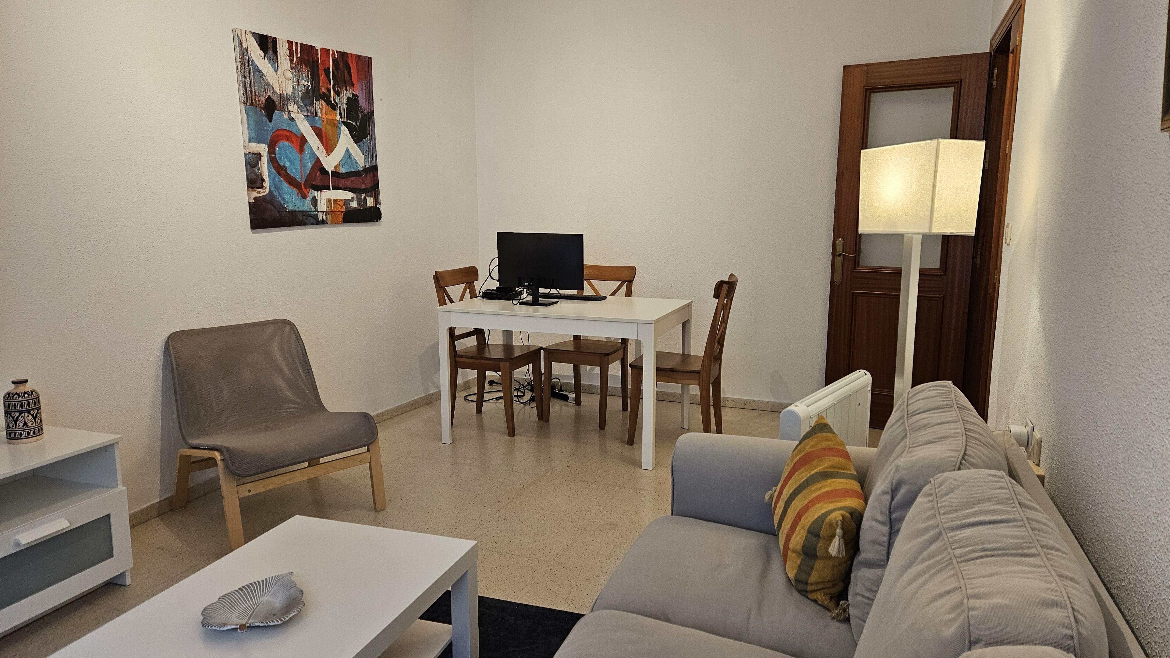Un salon avec une table blanche, un canapé gris, un fauteuil marron, une table à manger avec des chaises, un téléviseur et un tableau accroché au mur.