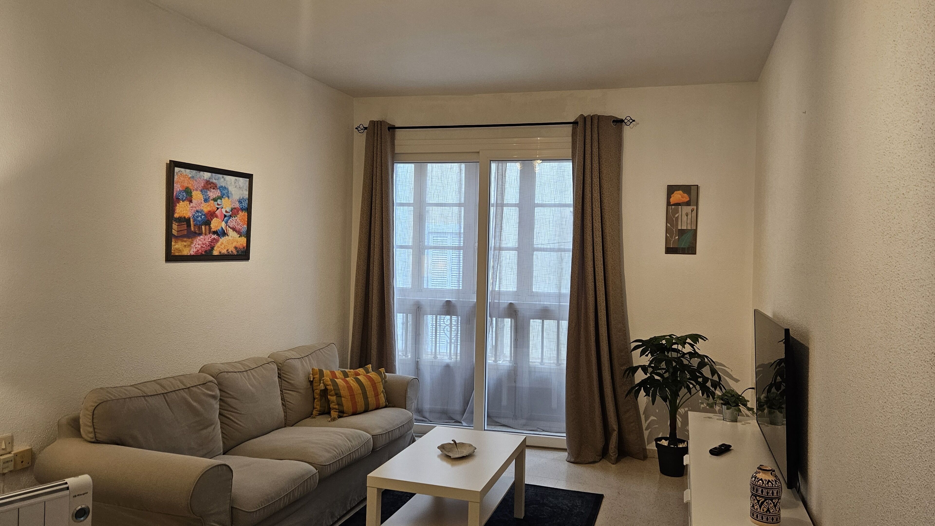 Un salon avec un canapé beige, une petite table blanche, une plante en pot et une fenêtre avec des rideaux.