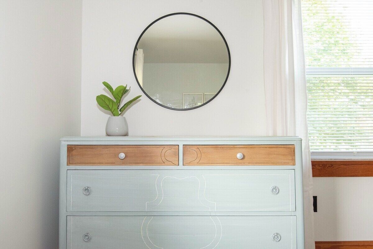 Une commode bleu clair avec des éléments en bois, un miroir rond et une petite plante.
