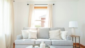 Un salon avec un canapé gris, une table d’appoint avec une lampe et une fenêtre avec des rideaux.