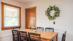 Une salle à manger avec une table et des chaises en bois, une fenêtre avec des stores, une porte et une guirlande accrochée au mur.