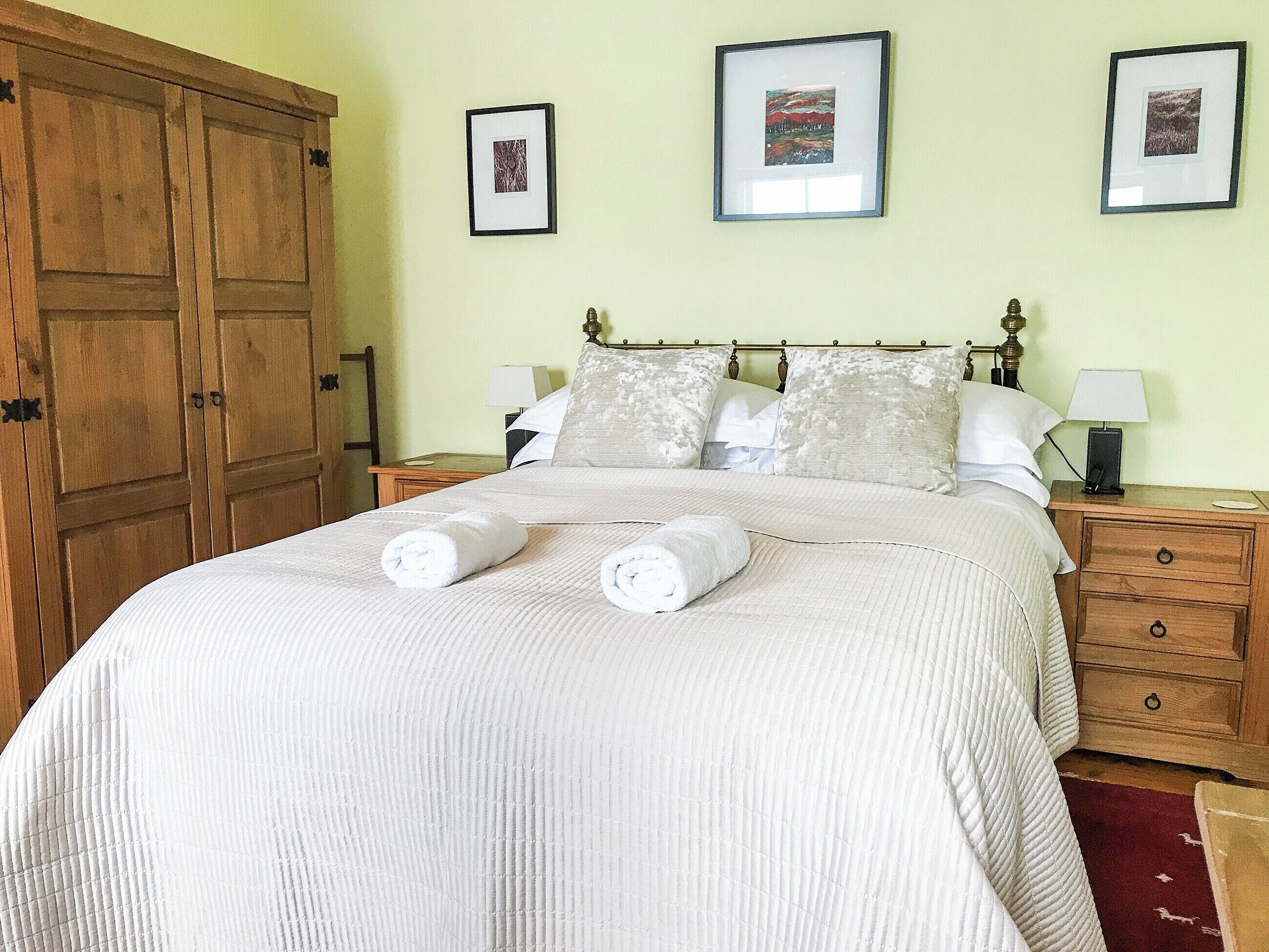 A neatly made bed with white linens and two rolled towels, a wooden wardrobe, a nightstand with a lamp, and two framed pictures on the wall.