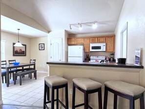 Cocina con armarios de madera, un refrigerador blanco, un microondas y una estufa. Hay tres taburetes de bar frente a la encimera.