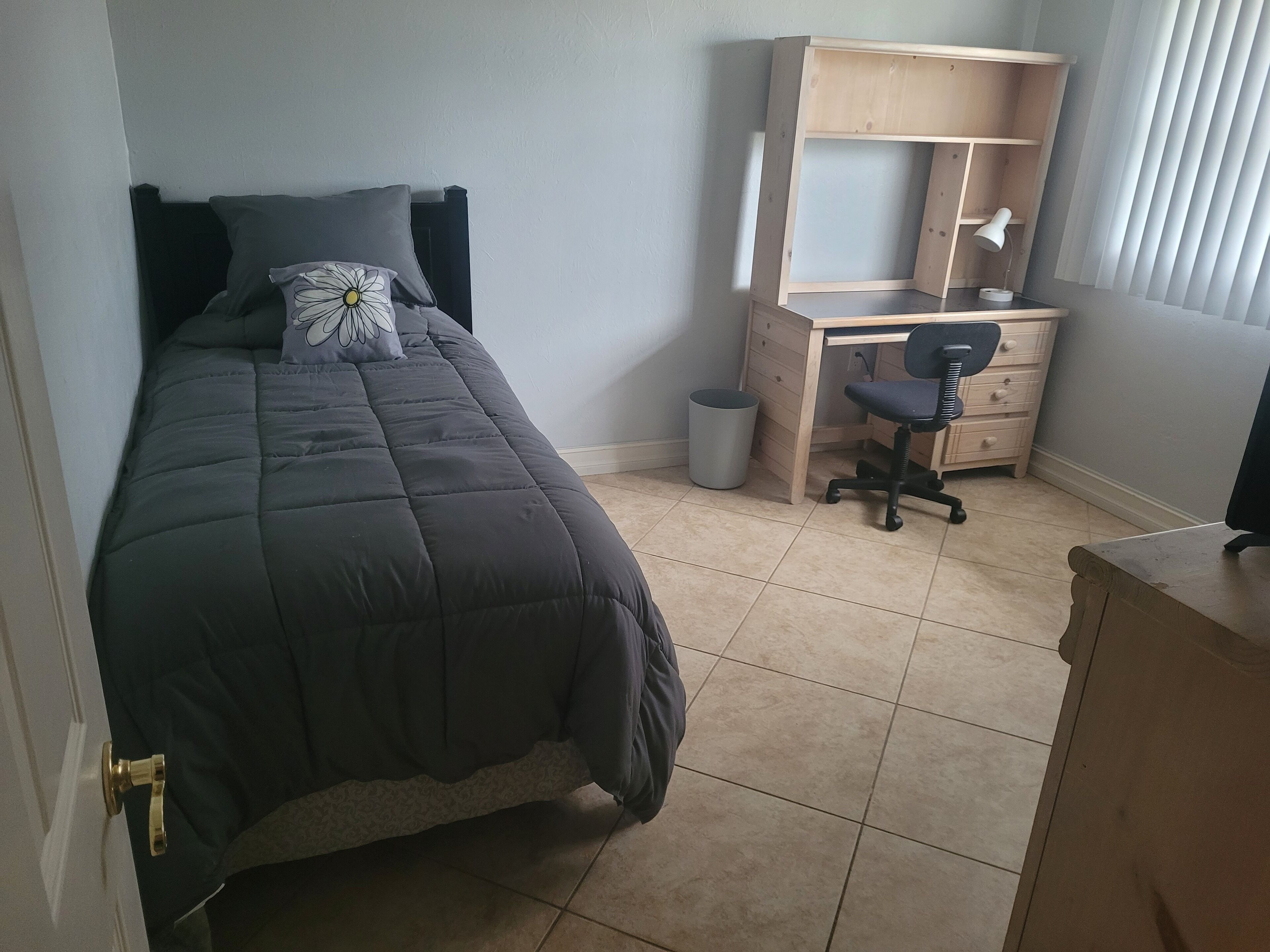 Ein Schlafzimmer mit Bett, Schreibtisch, Stuhl, Mülleimer und einem Fenster mit Jalousien.