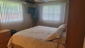Une chambre à coucher avec un lit, une commode en bois, un ventilateur de plafond et un téléviseur fixé au mur.