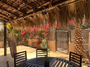 Eine Terrasse im Freien mit einem Reetdach, einem runden Tisch mit Stühlen und einer Wand mit roten Blüten.