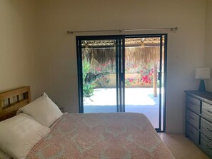 Ein Schlafzimmer mit einem Bett, einem Nachttisch, einer Kommode und einer Glastür, die zu einem Außenbereich mit einem Reetdach und Pflanzen führt.