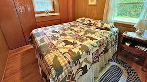 Ein Schlafzimmer mit einer holzverkleideten Wand, einem Bett mit gestecktem Bezug, einem Fenster mit Spitzenvorhängen und einem kleinen Nachttisch mit Lampe.