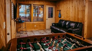 Un cuarto acogedor con paredes de madera, un sofá de cuero, una mesa de fútbol y una ventana con vista a un área boscosa.