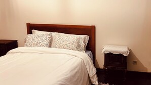 A bed with a wooden headboard, two pillows with floral patterns, a white bedspread, a striped rug, and a small bedside table with a white cloth. - Mom Home-007 (caldas da Rainha)