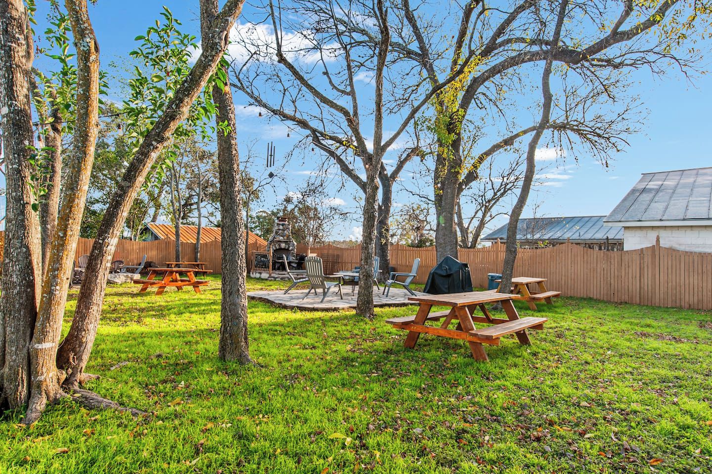 An outdoor patio area with picnic tables and a barbecue grill, surrounded by trees and a wooden fence.