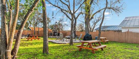 An outdoor patio area with picnic tables and a barbecue grill, surrounded by trees and a wooden fence.