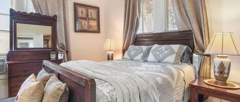 A bedroom with a wooden bed, a dresser, a mirror, a bedside table with a lamp, and a window with curtains.