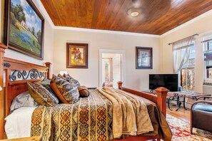 A bedroom with a wooden ceiling, a bed with patterned bedding, a television, and a window with curtains.