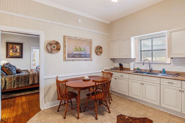 A dining area with a wooden table and chairs, a kitchen with white cabinets, and a window with blinds. - Carriage Inn - Bandera Suite 2 (Bandera)