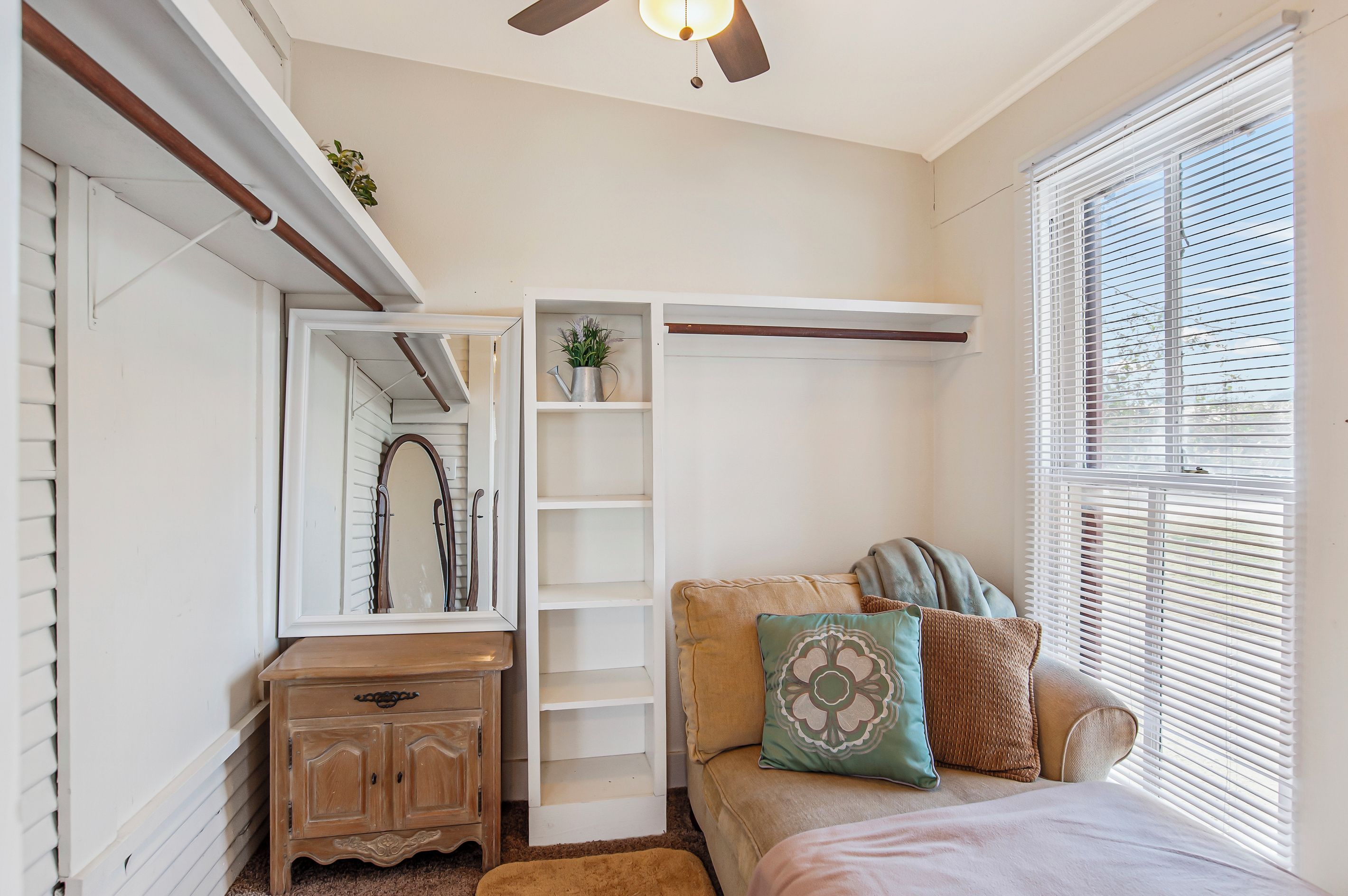 A bedroom with a bed, a dresser, a mirror, a shelf with a plant, and a window with blinds.
