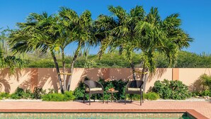 A pool area with two wicker chairs and a small table, surrounded by palm trees and a low brick wall.
