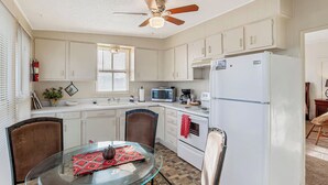 Una cocina con armarios blancos, microondas, refrigerador, ventilador de techo y una mesa de comedor con sillas.