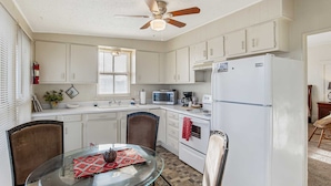 A compact kitchen with white cabinets, a microwave, a stove, and a refrigerator. There is a ceiling fan, a window with blinds, and a glass table with chairs. - Carriage Inn - Tarpley Suite 5 (Bandera)