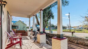 A covered porch with stone flooring, red and black outdoor furniture, and a view of a residential area with houses and a road. - Carriage Tarpley 5 | Queen Suite w/ Kitchen (Bandera)
