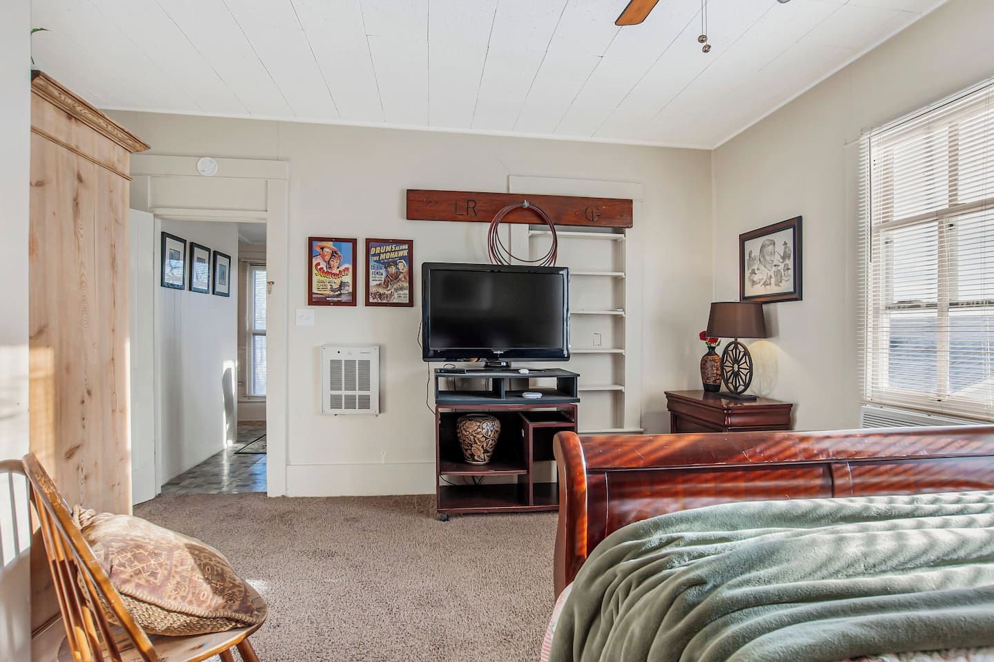 A bedroom with a bed, a TV, a wooden cabinet, and a window with blinds.