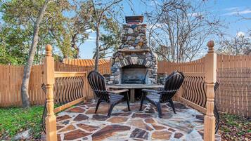 An outdoor stone patio with a stone fireplace, two chairs, and a table.