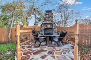 An outdoor stone patio with a stone fireplace, two chairs, and a table.