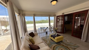 Amplia sala de estar con un sofá color beige, un otoman amarillo y una alfombra estampada. Hay un comedor con mesa y sillas, y vistas a la playa a través de grandes ventanales.