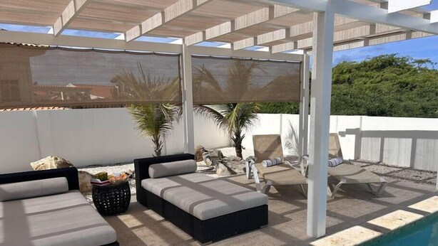 Zona junto a la piscina con un espacio de descanso cubierto, una piscina y una valla blanca.
