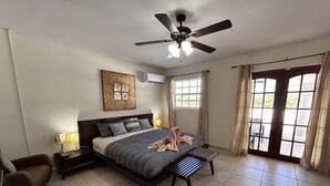 Un dormitorio con cama, lámparas de noche, ventilador de techo, ventana con cortinas y una obra de arte colgada en la pared.
