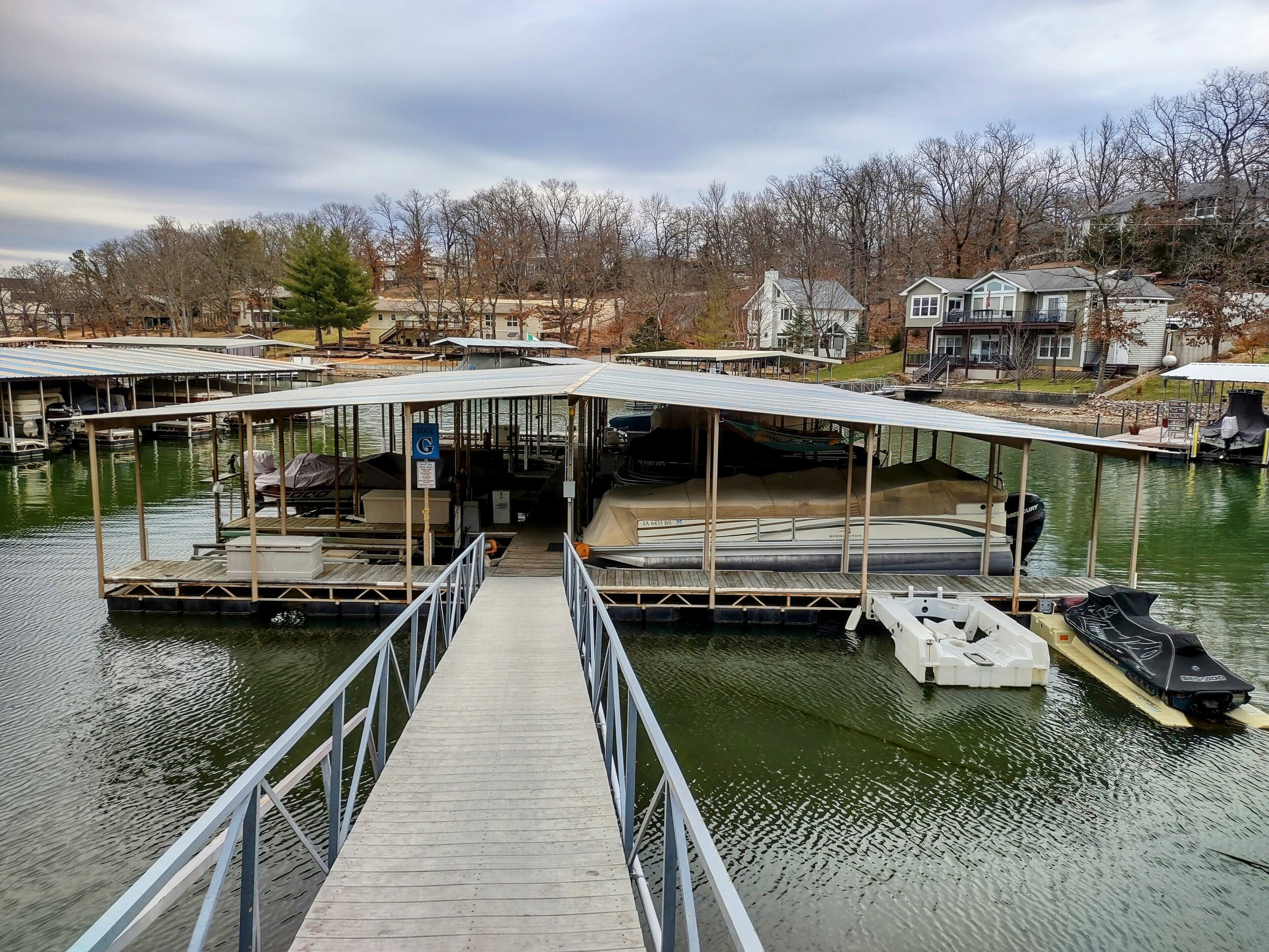 A dock with a covered walkway leading to a boat launch area, surrounded by houses and trees.