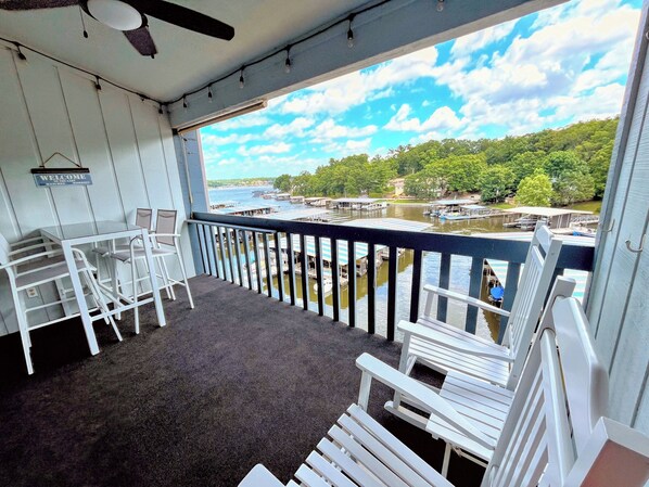 A balcony with white chairs and a table, overlooking a river with boats and docks.