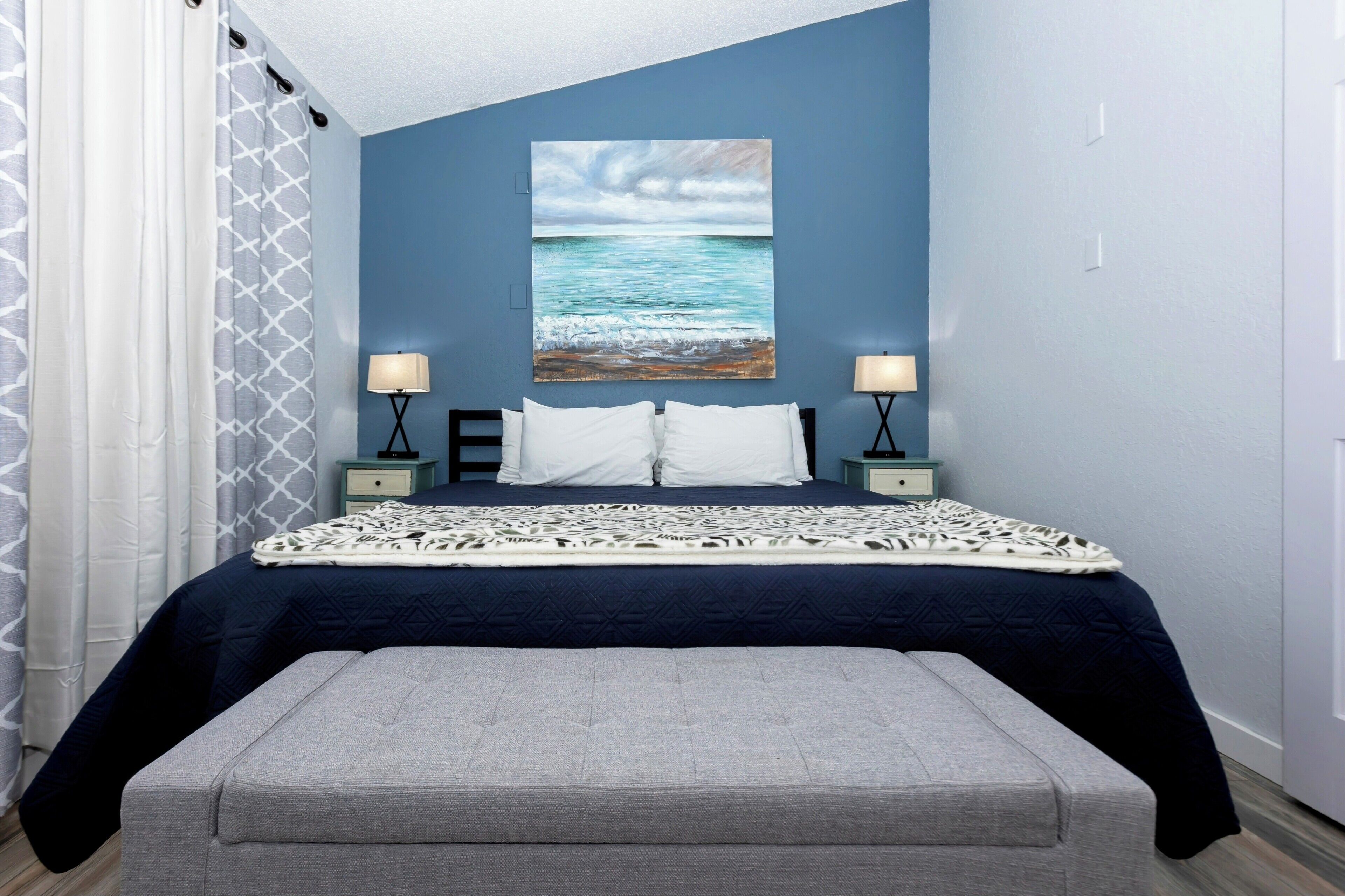 A bedroom with a bed, two bedside tables, a bench, a painting of the ocean, and a window with curtains.