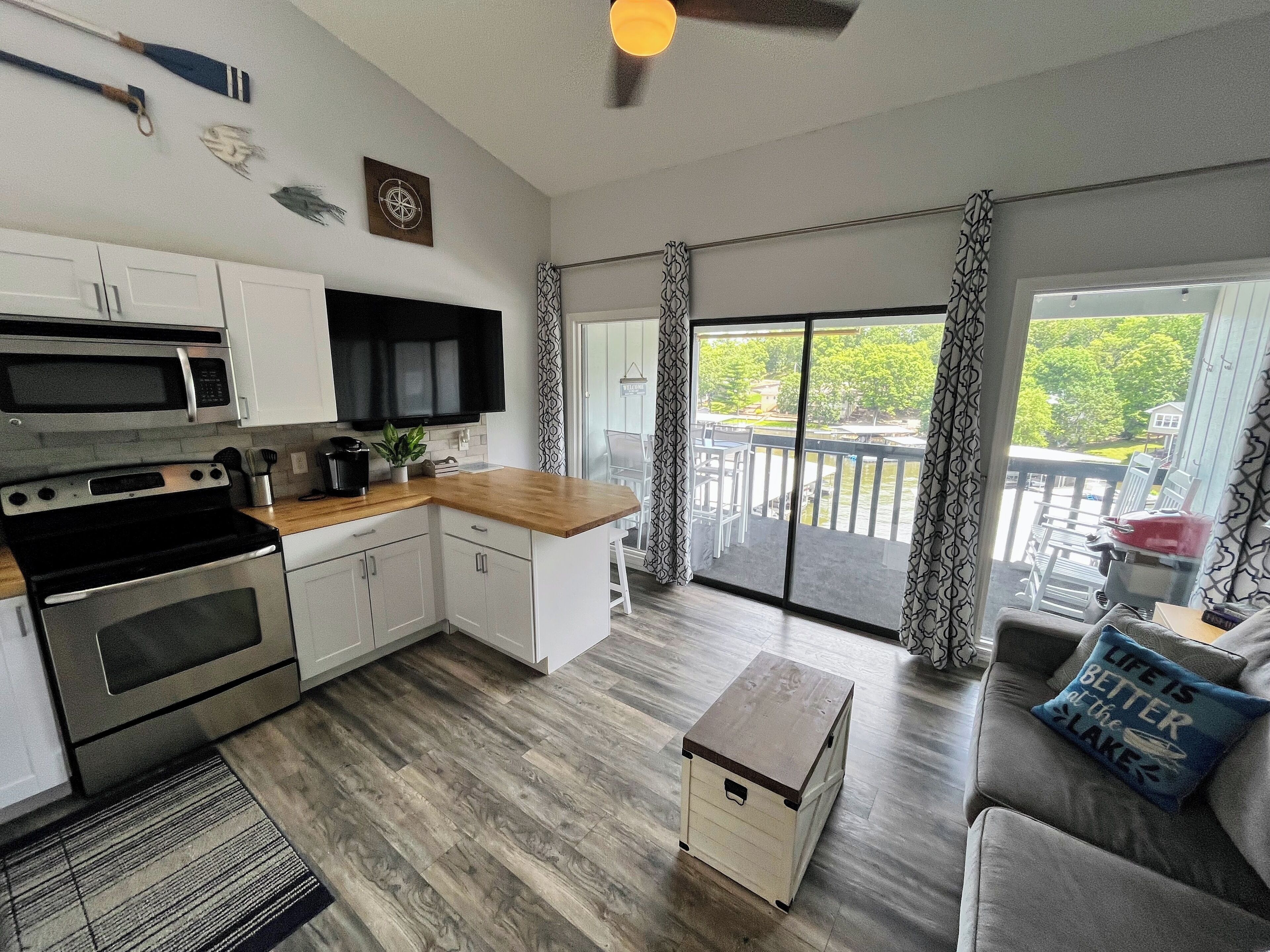 A modern kitchen with white cabinets, a microwave, and an oven. There is a wooden countertop, a small plant, and a coffee maker. A sliding glass door leads to a balcony with outdoor furniture.