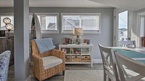 Una habitación con una silla de mimbre, una consola blanca con libros, una mesa de comedor con sillas y ventanas amplias.