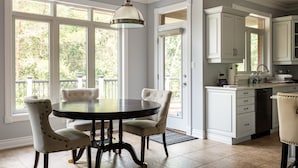 Une cuisine avec des meubles blancs, une table à manger avec des chaises et une grande fenêtre donnant sur les arbres.