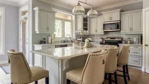 Une cuisine spacieuse avec des meubles blancs, un grand îlot central doté de tabourets, un lustre et un four à micro-ondes.