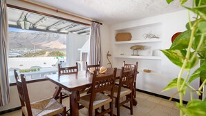 A dining area with a wooden table and chairs, a view of mountains through a window, and a shelf with decorative items. - Caracolillos House, Famara Surf Beach (Famara)