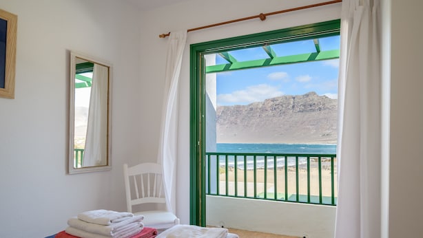 A room with a bed, a chair, and a balcony with a view of the beach and mountains. - Caracolillos House, Famara Surf Beach (Famara)