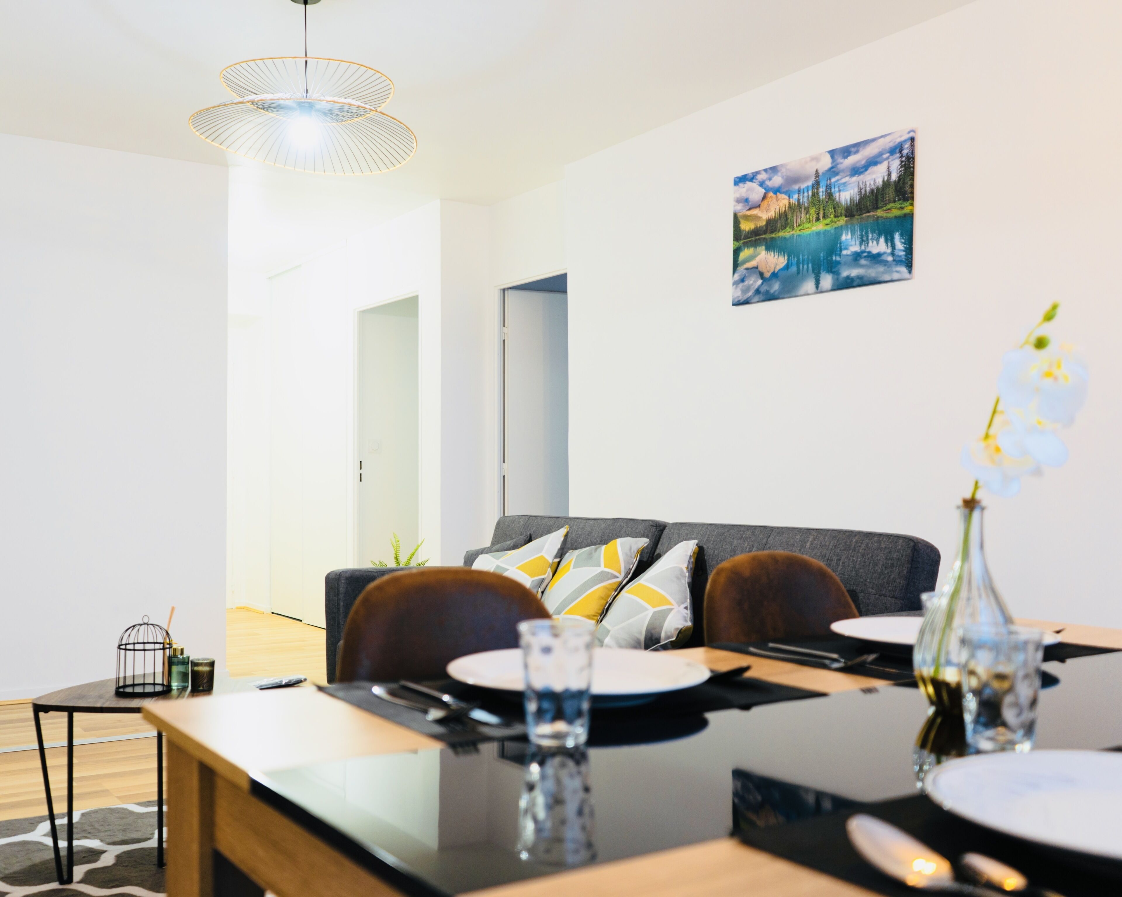 Ein moderner Essbereich mit einem Holztisch, gedeckt für zwei Personen, einer grauen Couch mit gemusterten Kissen und einem Landschaftsbild an der Wand.