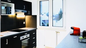 Une cuisine moderne avec des meubles de couleur noire, un four encastré, un four à micro-ondes et une machine à café rouge. Une fenêtre offre une vue sur les maisons à l’extérieur.