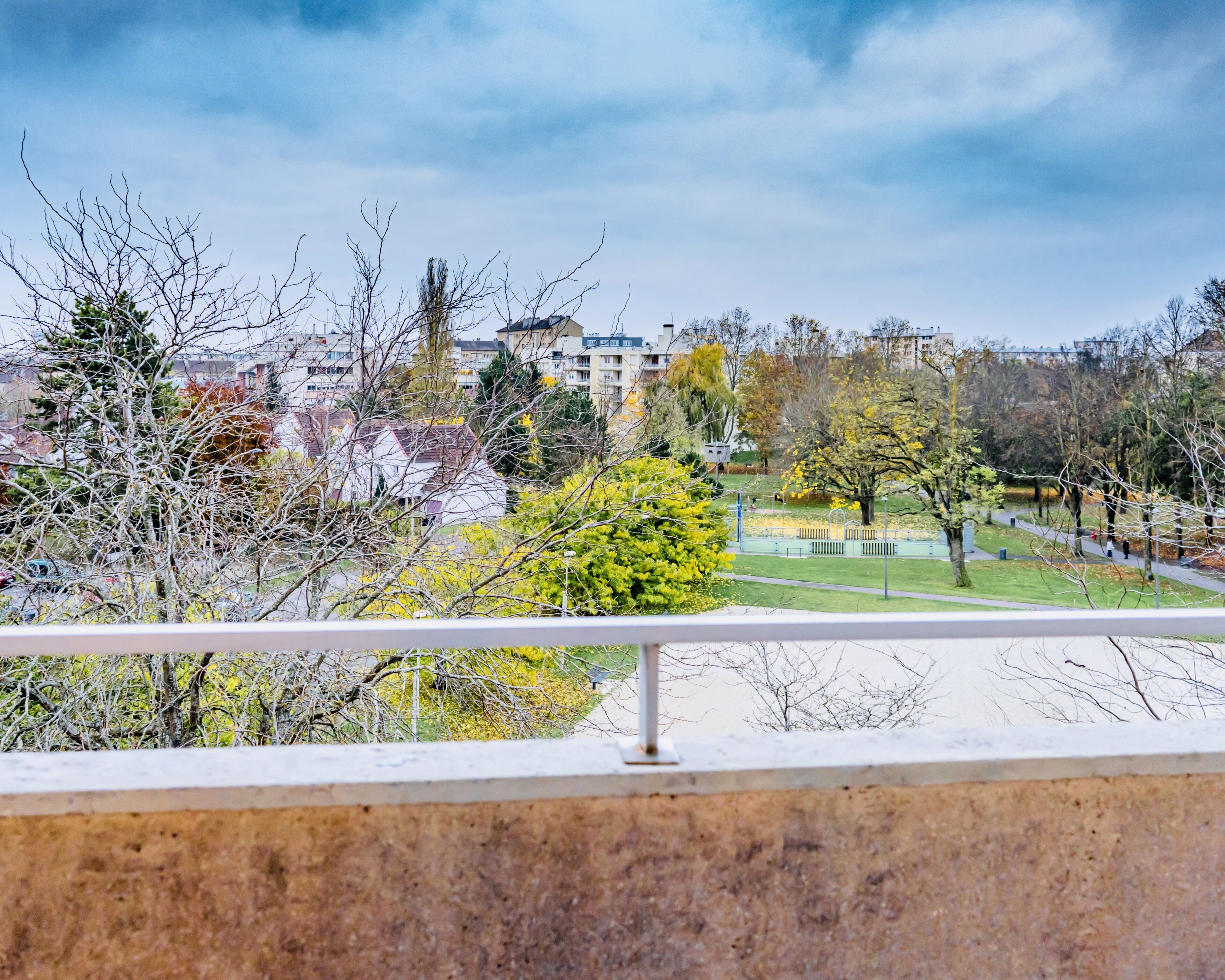 Vista desde un balcón sobre un parque con árboles y edificios al fondo.