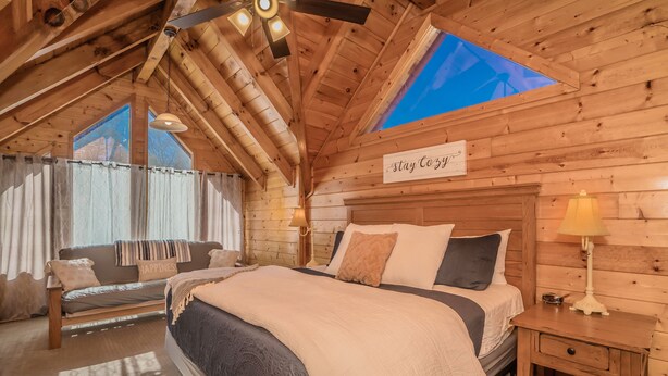 A cozy log cabin bedroom with a large bed, a sofa, a small table, and a ceiling fan.