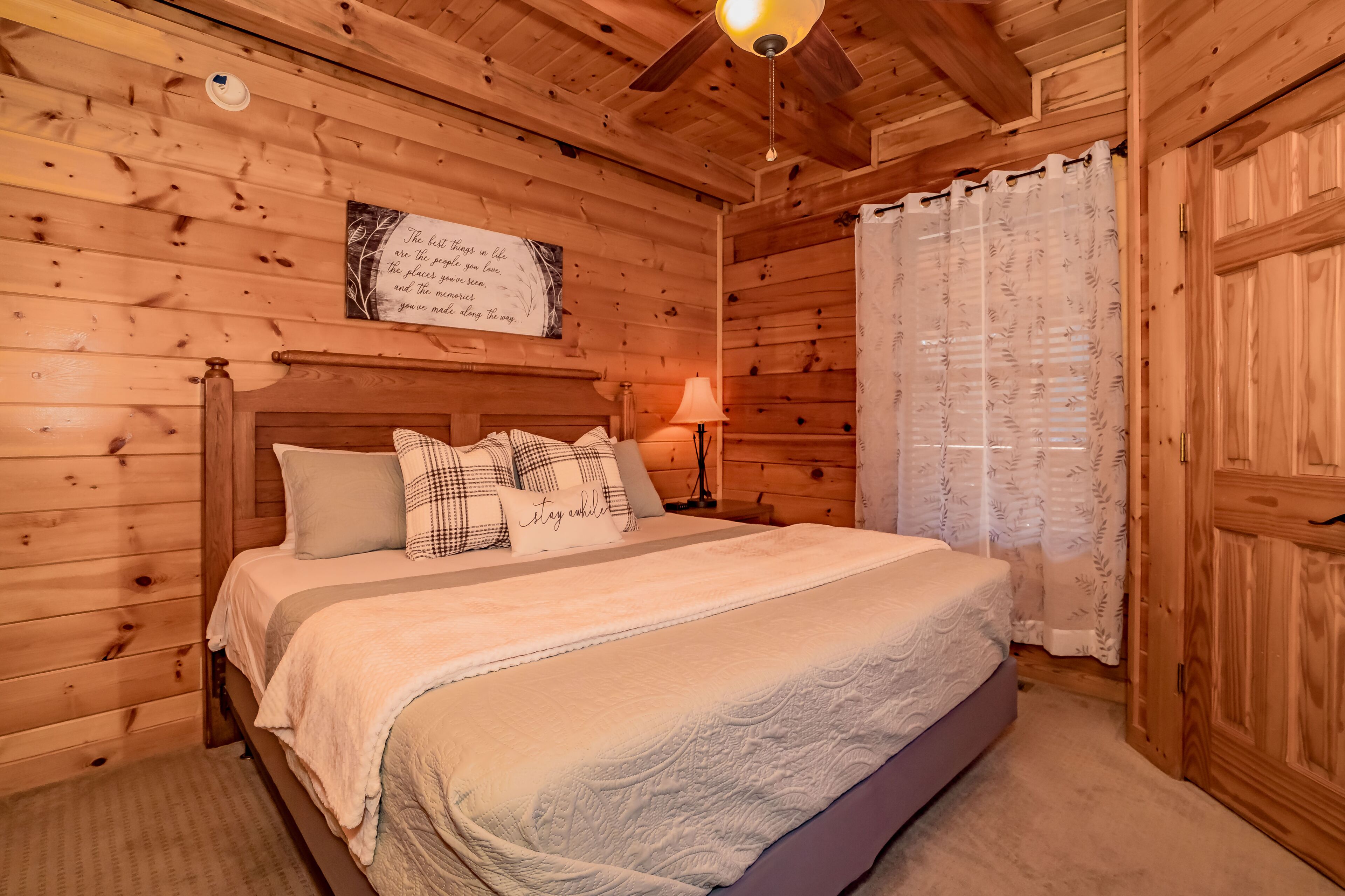 A wooden cabin bedroom with a bed, a ceiling fan, a nightstand, and a wall-mounted sign.