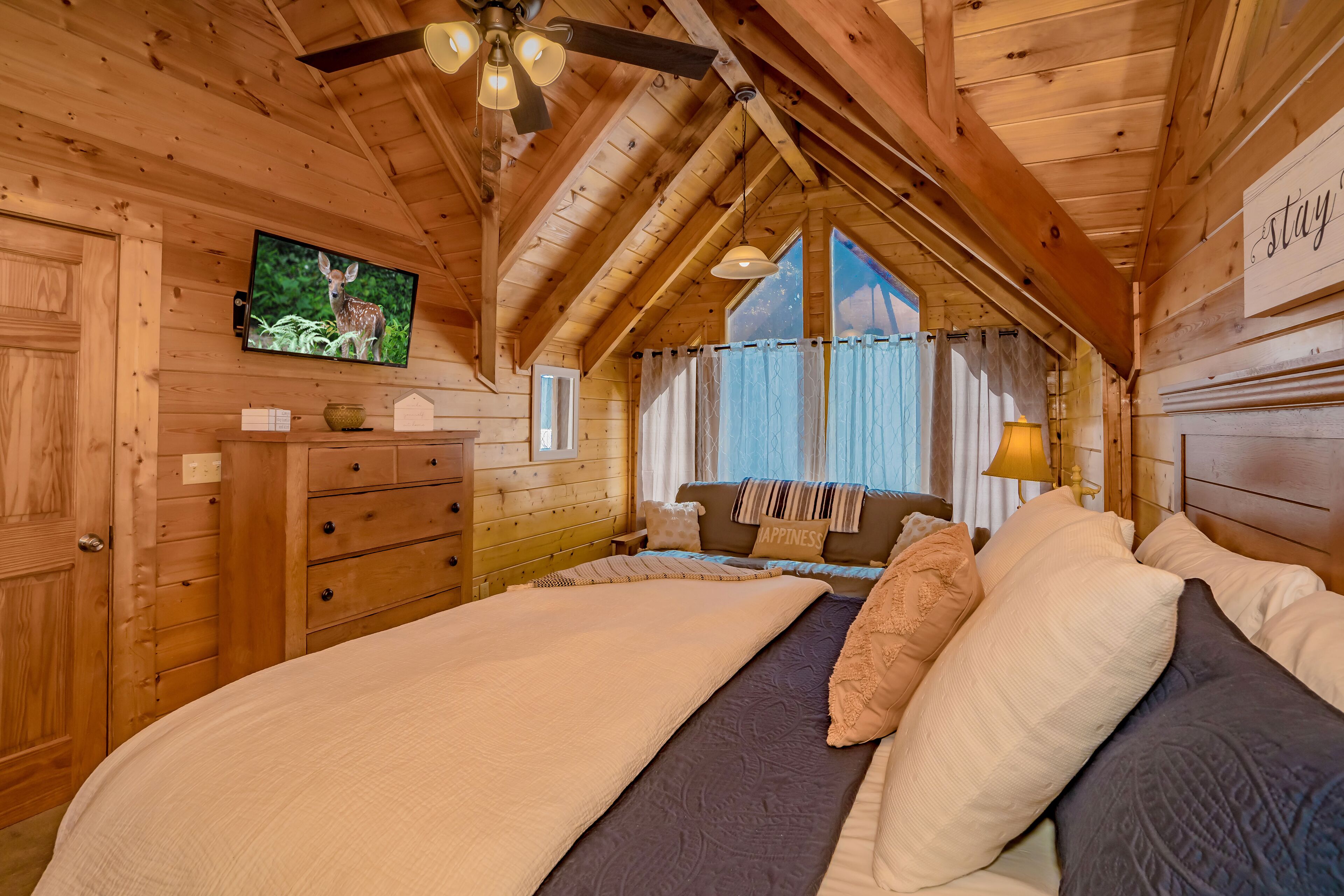 Un dormitorio rústico de cabaña con cama, cómoda, televisor y ventana con cortinas.