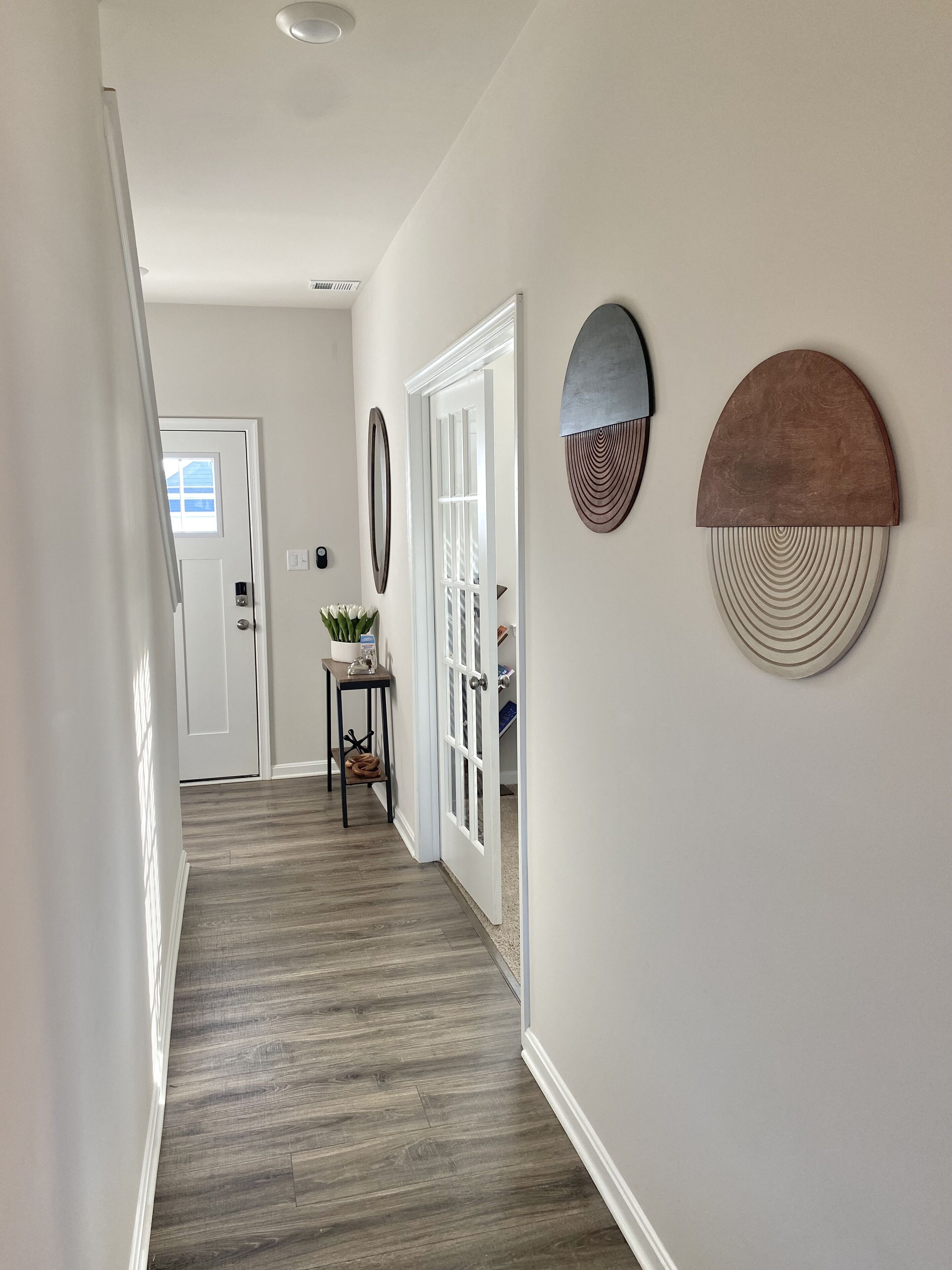 Un couloir doté d’un plancher en bois, de murs blancs et d’une porte donnant sur une autre pièce. On y trouve deux objets décoratifs fixés au mur et une petite table sur laquelle est posée une plante.