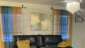 A living room with a black leather sofa, two yellow throw pillows, a glass coffee table, and a ceiling fan with a crystal chandelier.
